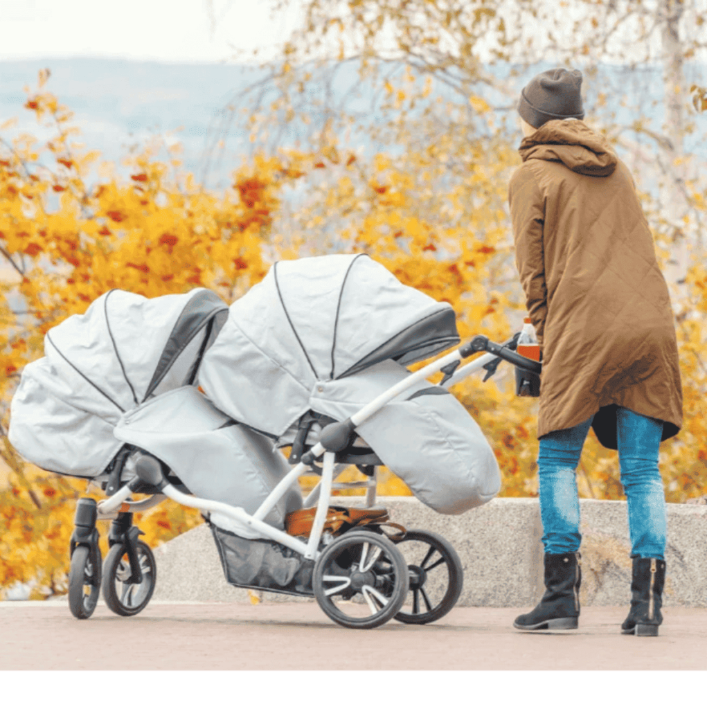 Mãe empurrando seu carrinho duplo de bebês gêmeos, grande e na cor cinza claro, e ao fundo um lindo jardim de flores amarelas. Os Melhores Carrinhos para Bebês Gêmeos