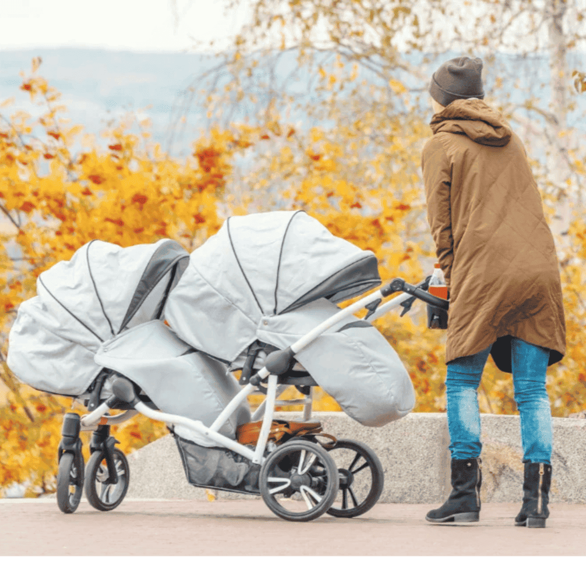 Mãe empurrando seu carrinho duplo de bebês gêmeos, grande e na cor cinza claro, e ao fundo um lindo jardim de flores amarelas. Os Melhores Carrinhos para Bebês Gêmeos