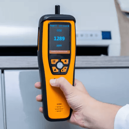 Mão masculina segurando um medidor de qualidade do ar próximo à saída de ar de um ar condicionado, em um ambiente interno. Os Melhores Medidores de Qualidade do Ar