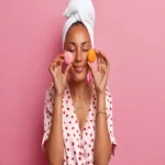 Mulher com toalha na cabeça e roupão de poá rosa segurando dois macarons (rosa e laranja) sobre os olhos, simbolizando dicas e reviews de produtos de cuidado pessoal, beleza e bem-estar.