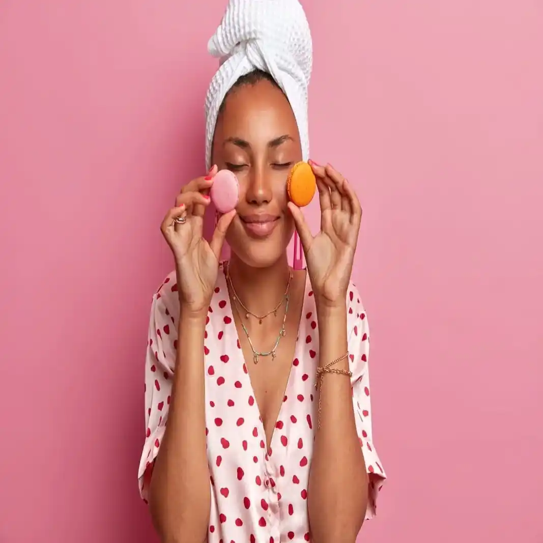 Mulher com toalha na cabeça e roupão de poá rosa segurando dois macarons (rosa e laranja) sobre os olhos, simbolizando dicas e reviews de produtos de cuidado pessoal, beleza e bem-estar.