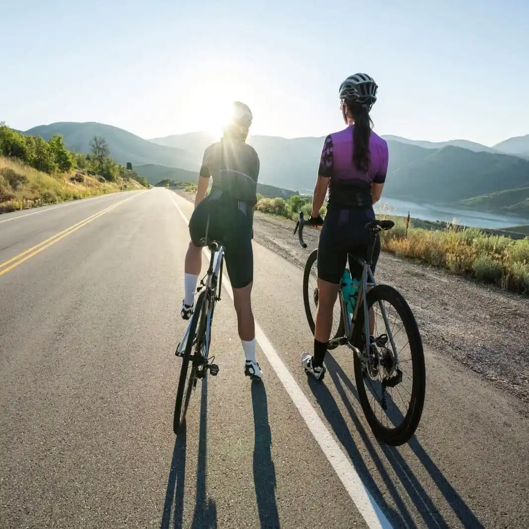 Vista traseira de dois ciclistas de estrada, vestindo roupas de ciclismo e capacetes, parados com suas bicicletas em uma estrada de asfalto nas montanhas durante o pôr do sol, representando equipamentos de esporte e mobilidade.