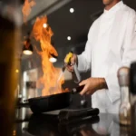 Chef de cozinha profissional em dolmã branca segurando uma frigideira preta que emite grandes chamas azuis e laranjas ao usar um maçarico de cozinha, ilustrando equipamentos de gastronomia e culinária.