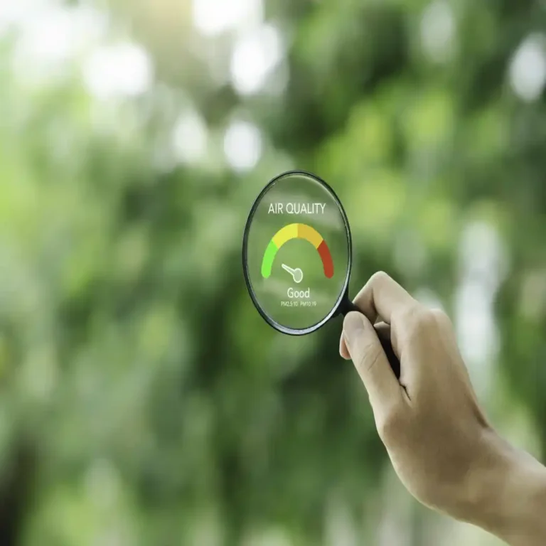 Mão segurando uma lupa sobre um fundo desfocado de natureza verde, focando em um gráfico digital de 'Qualidade do Ar' com ponteiro na zona verde 'Bom', representando medidores de ar e sensores ambientais.
