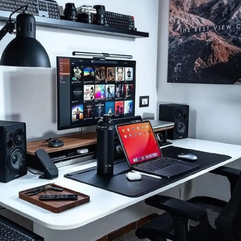 Setup de escritório moderno e organizado com mesa branca, monitor grande mostrando biblioteca de músicas, tablet com tela vermelha, alto-falantes pretos, luminária de mesa e acessórios, representando reviews de tecnologia e home office.