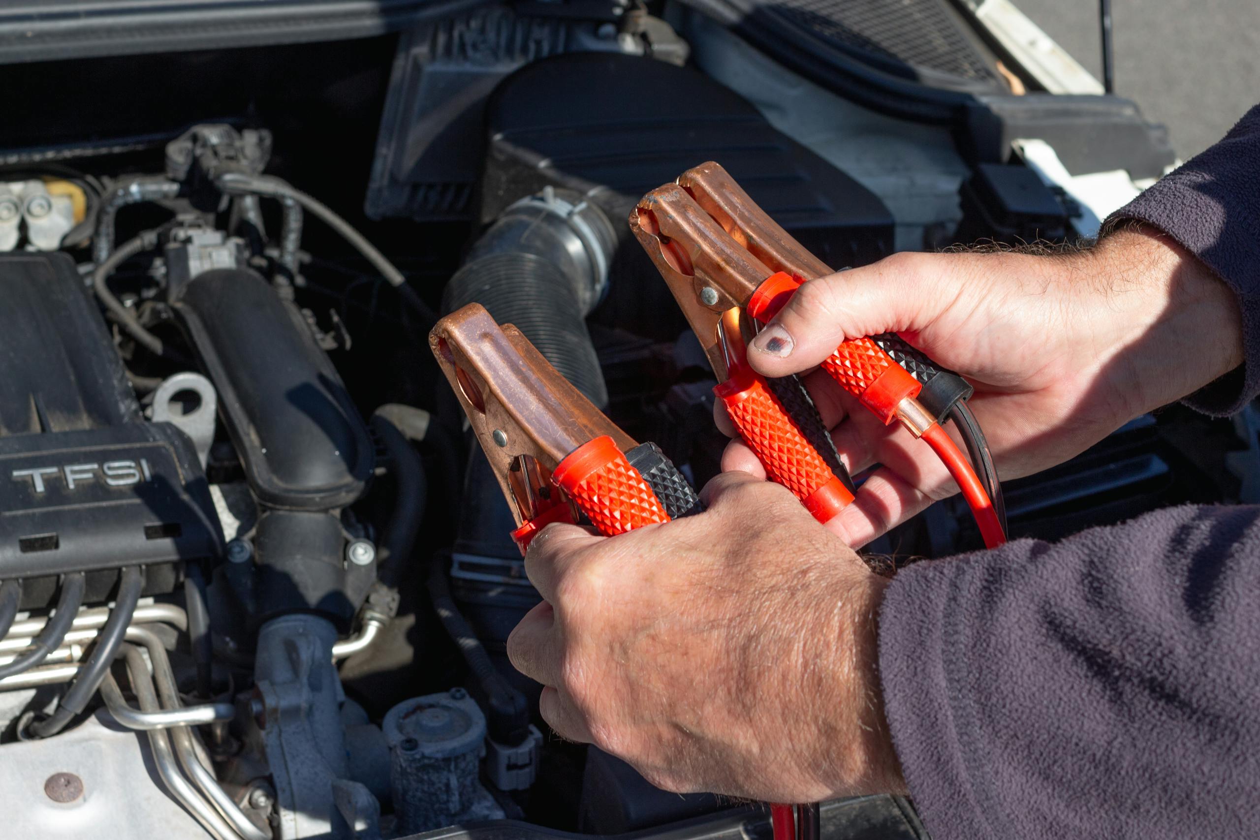 Melhor auxiliar de partida portátil. Imagem em close-up de mãos segurando cabos de ligação perto do motor de um carro, prontos para o conserto.
